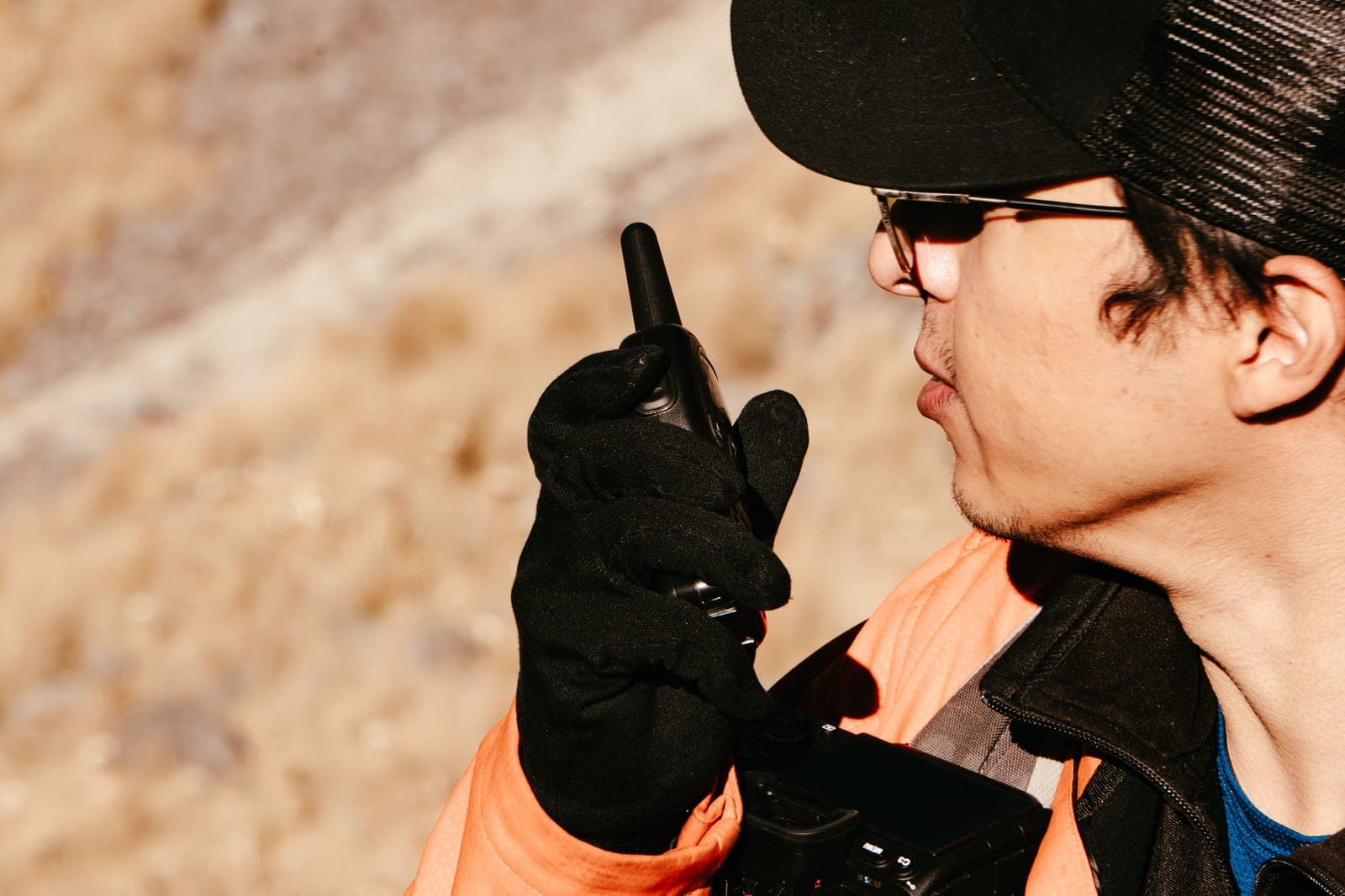 A man in a cap and gloves talking into a handheld radio outdoors